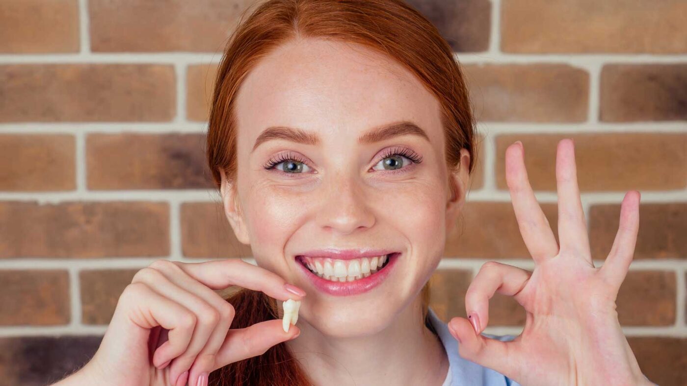 A smiling woman holding a tooth and making an OK gesture