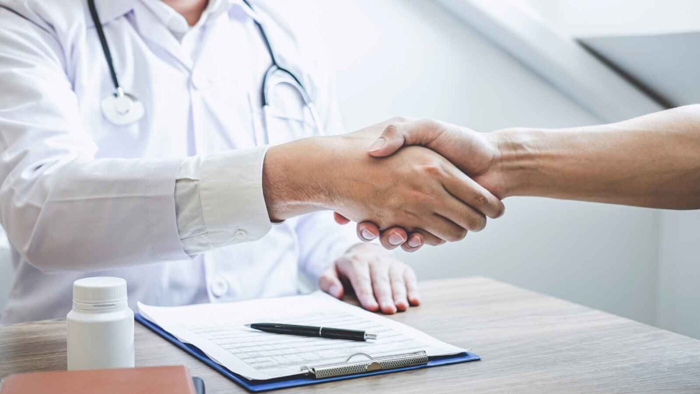 A doctor and a patient shake hands over a desk