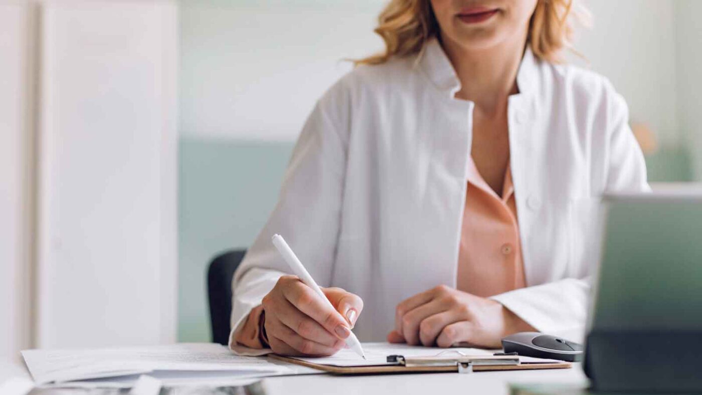 A female doctor in a white coat writes on a clipboard at her desk.