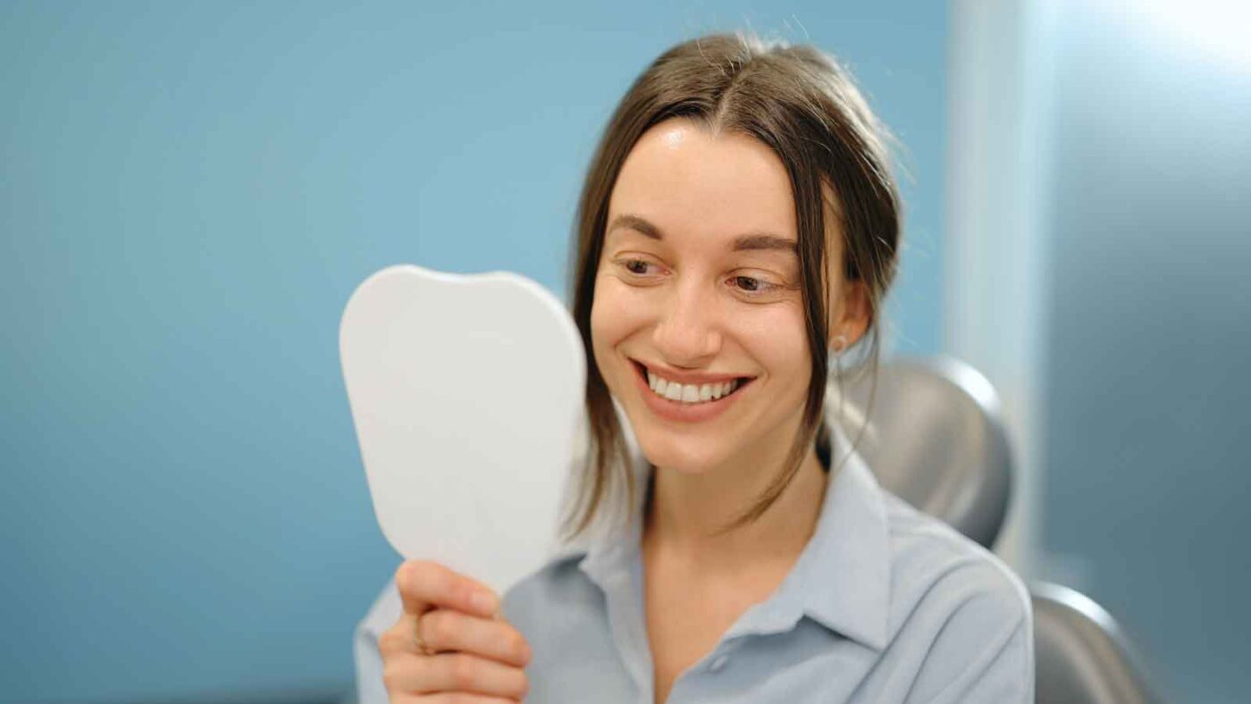 A smiling woman looks at her reflection in a handheld mirror.